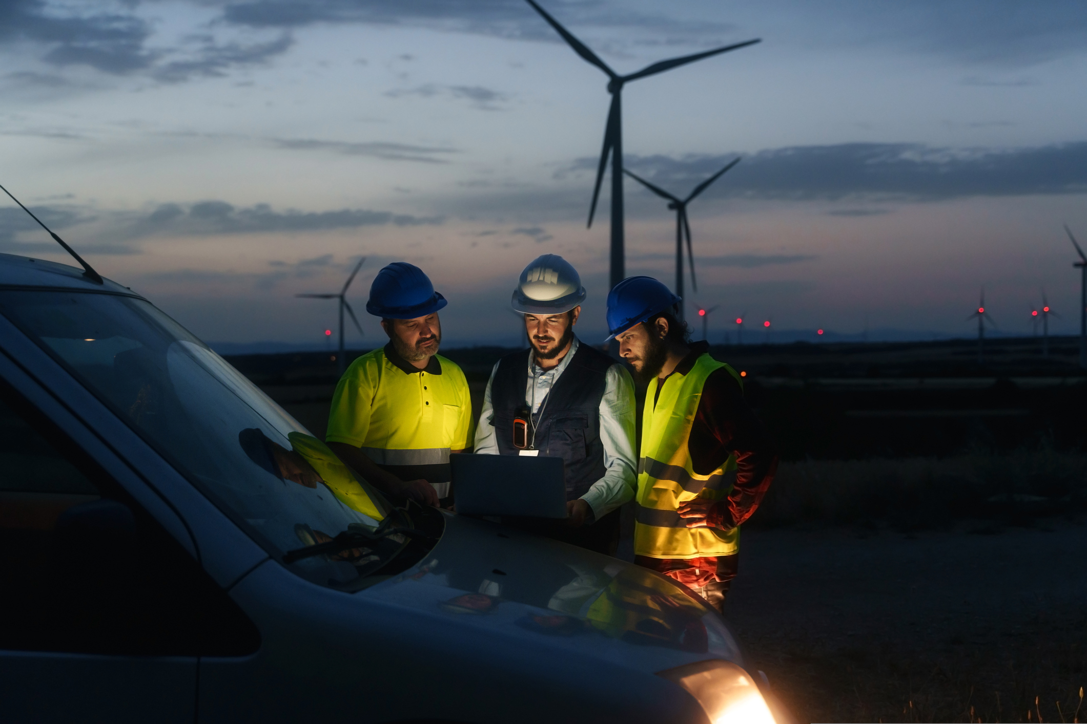 Engineers in safety vests and helmets discussing renewable energy solutions on a laptop at a wind turbine electricity plant during twilight