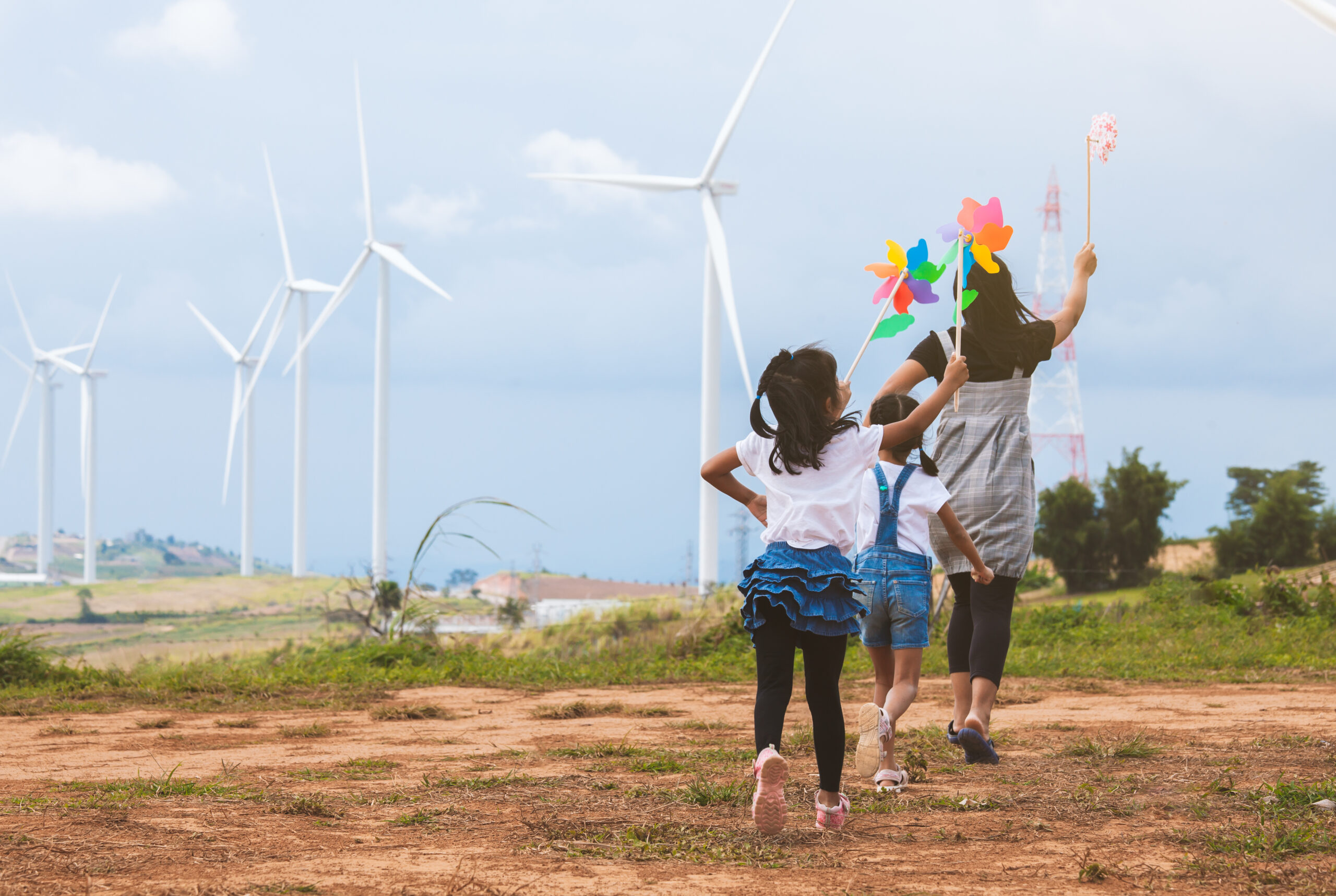 Back view of two children and an adult walking towards wind turbines, the adult holding a colourful pinwheel up in the air