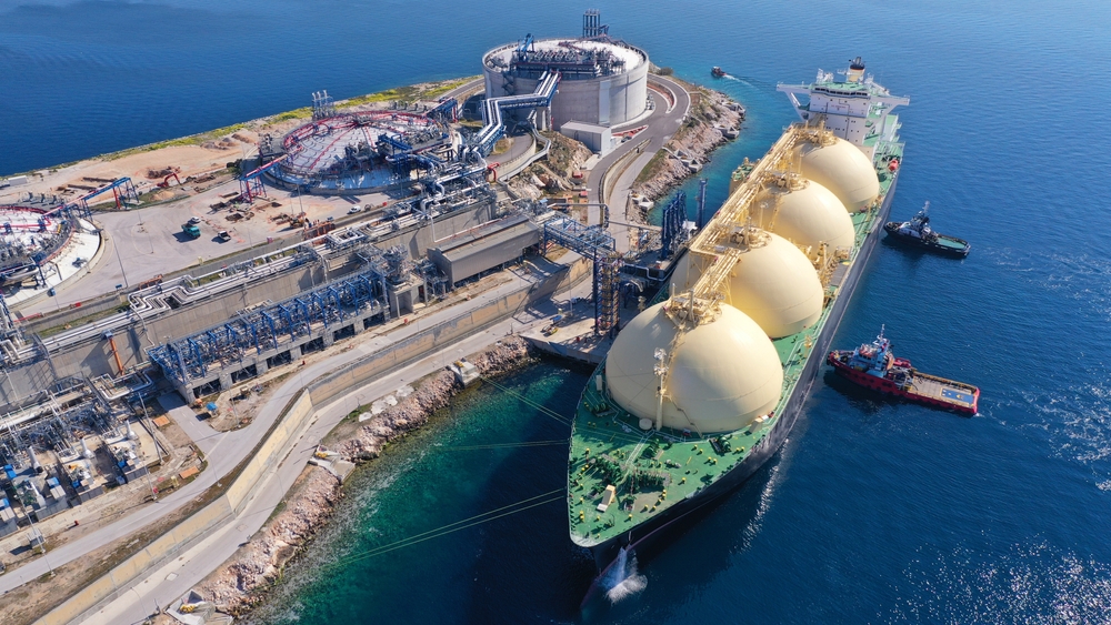 Aerial view of an LNG tanker docked at a coastal industrial facility with distinctive spherical storage tanks and infrastructure for natural gas.