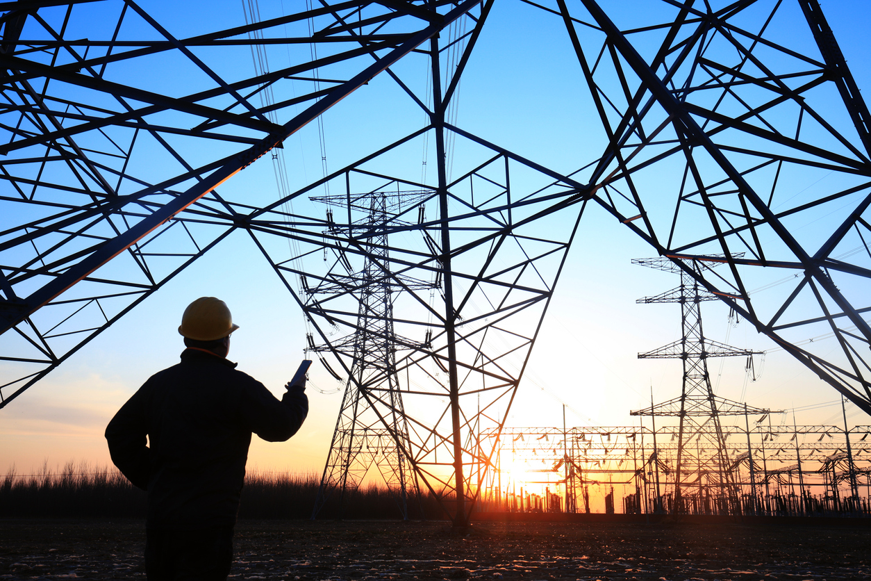 Safety worker in hard hat pointing at electrical transmission towers under a colorful sunset sky, highlighting energy infrastructure.