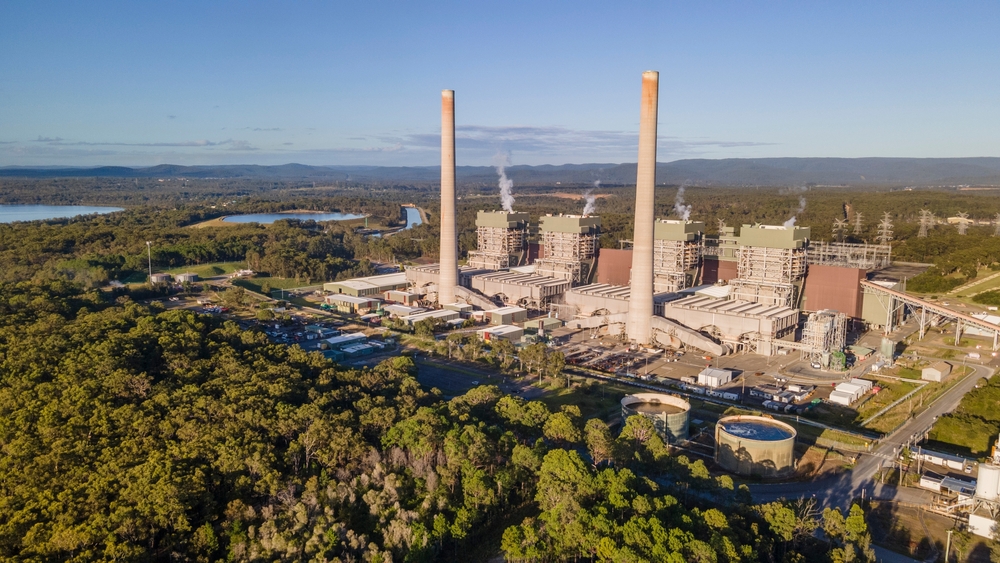 Aerial view of a coal-fired power station with tall chimneys emitting smoke, surrounded by forest and a body of water in the distance.
